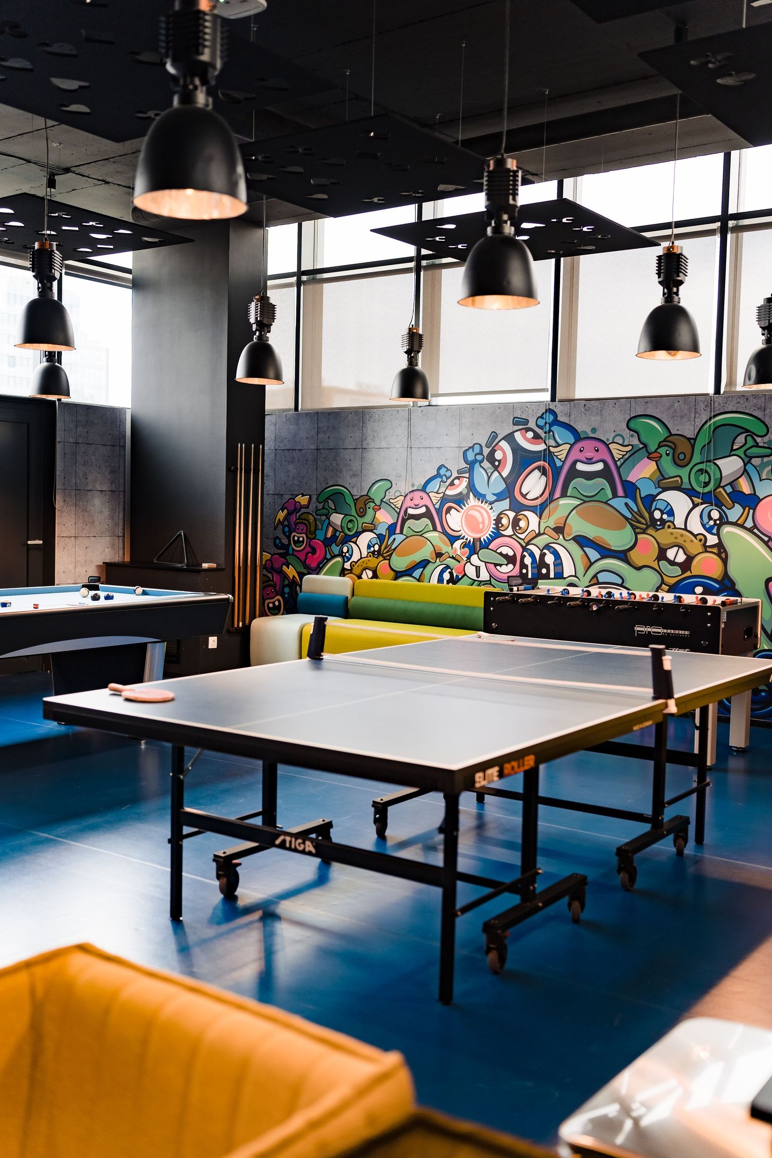 Office game room with a ping pong table, pool table, foosball and a colorful graffiti mural under black pendant lights.