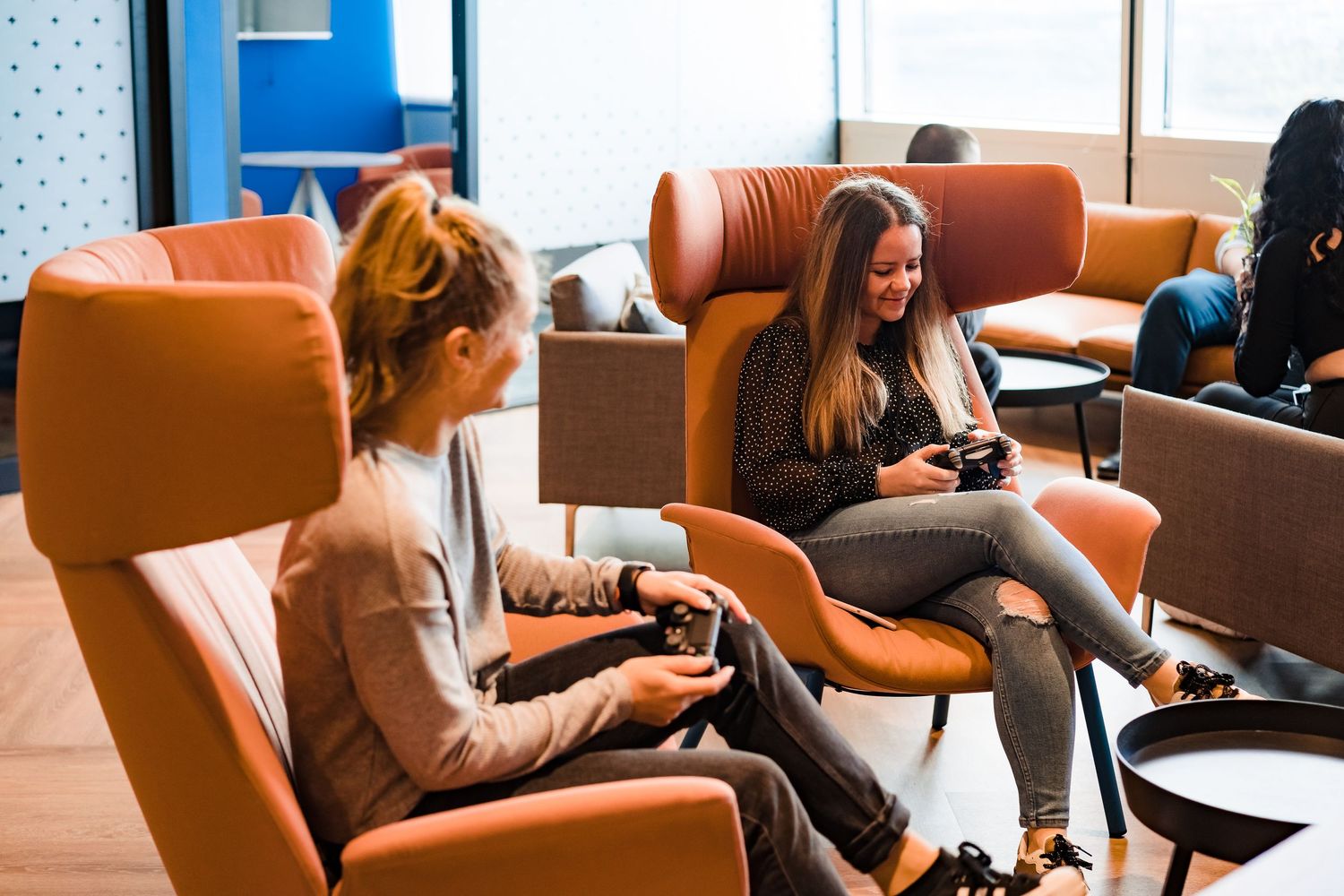 Two women relax in orange armchairs holding game controllers while playing video games in a bright office break area.