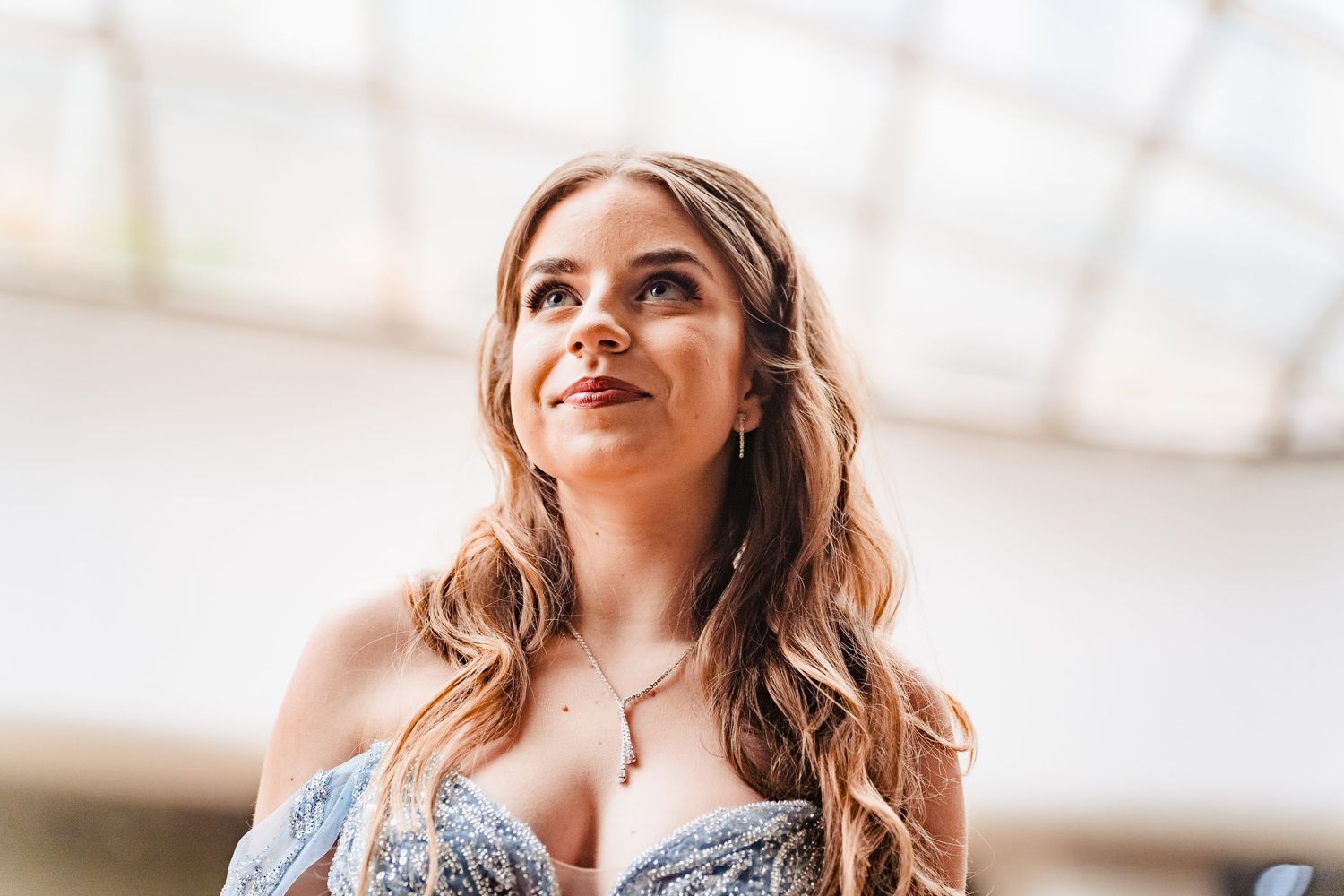 Close-up portrait of a smiling young woman with long wavy hair wearing a pendant necklace and a blue lace gown.
