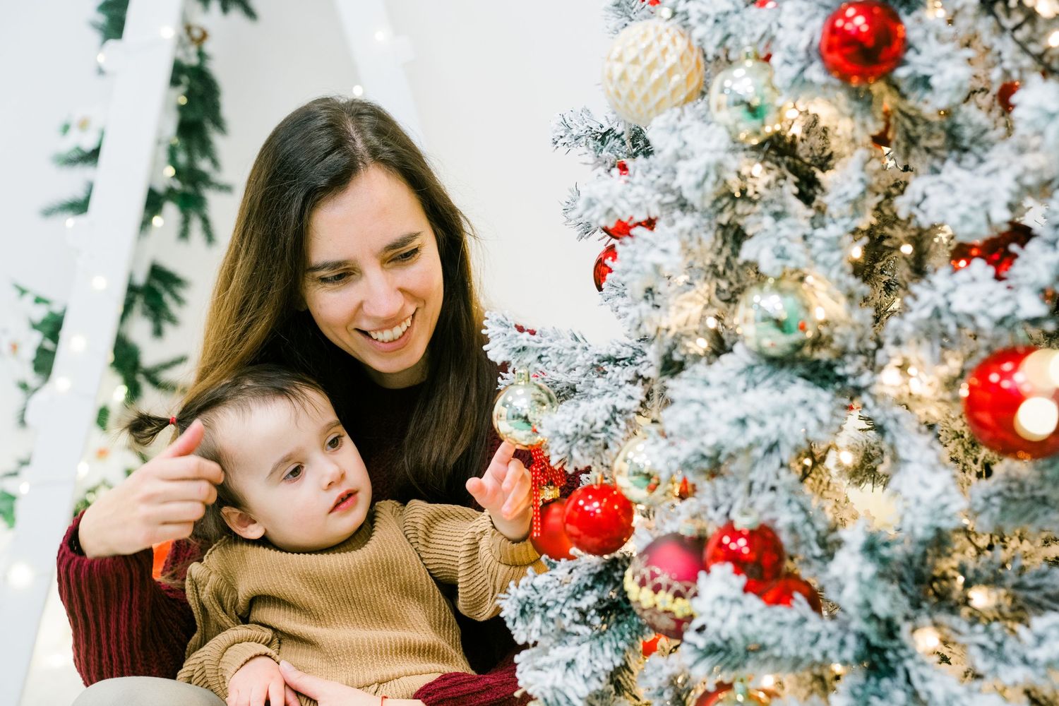 Smiling mother and toddler son admire the red and gold ornaments on a frosted Christmas tree.