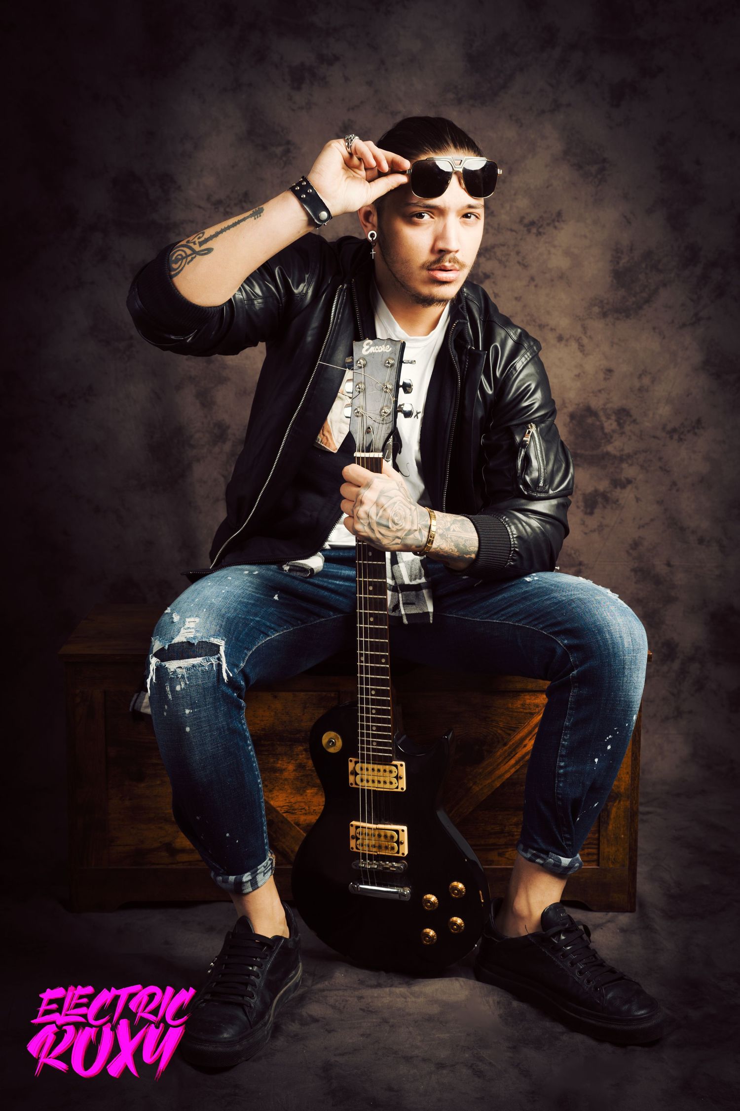 Tattooed guitarist in leather jacket and ripped jeans sits on a wooden crate, lifting his sunglasses while holding a black electric guitar.