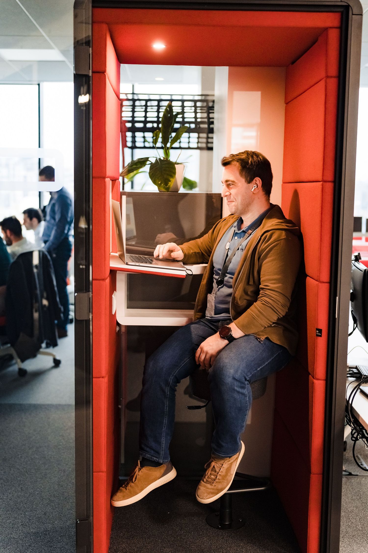 Smiling man in a brown jacket sits on a stool in a red office pod, working on a laptop in a modern open workspace.