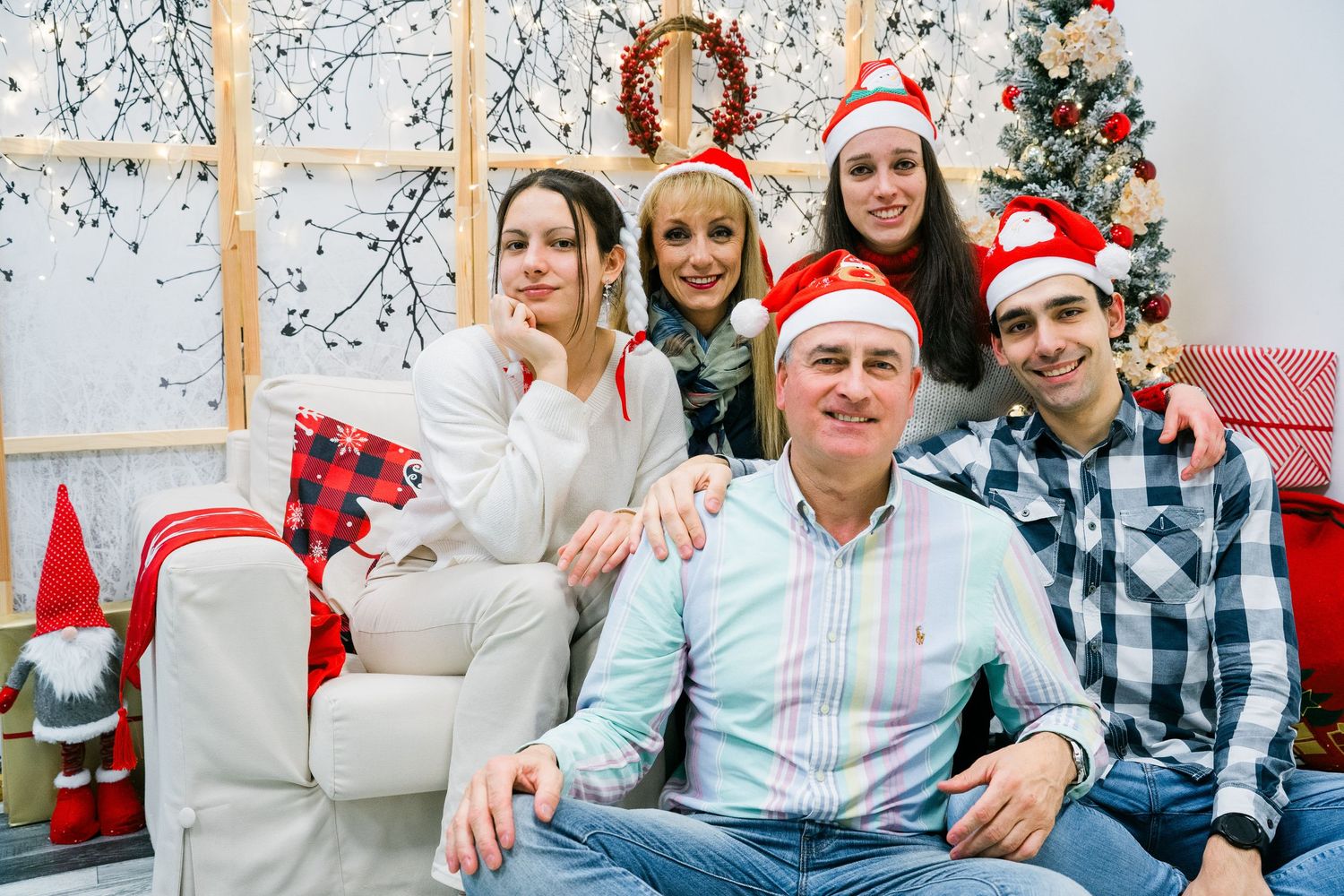 Five smiling family members in Santa hats pose together on a white sofa near a decorated Christmas tree.