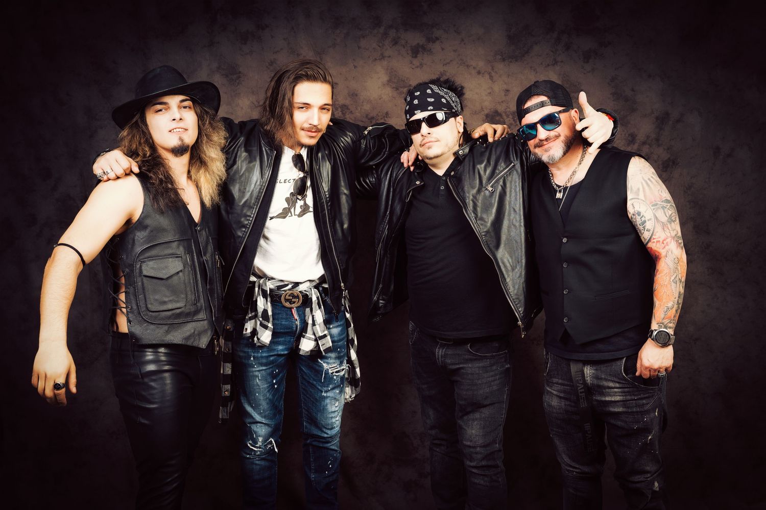 Four rock musicians in leather jackets and sunglasses stand with arms around each other against a dark moody studio backdrop.