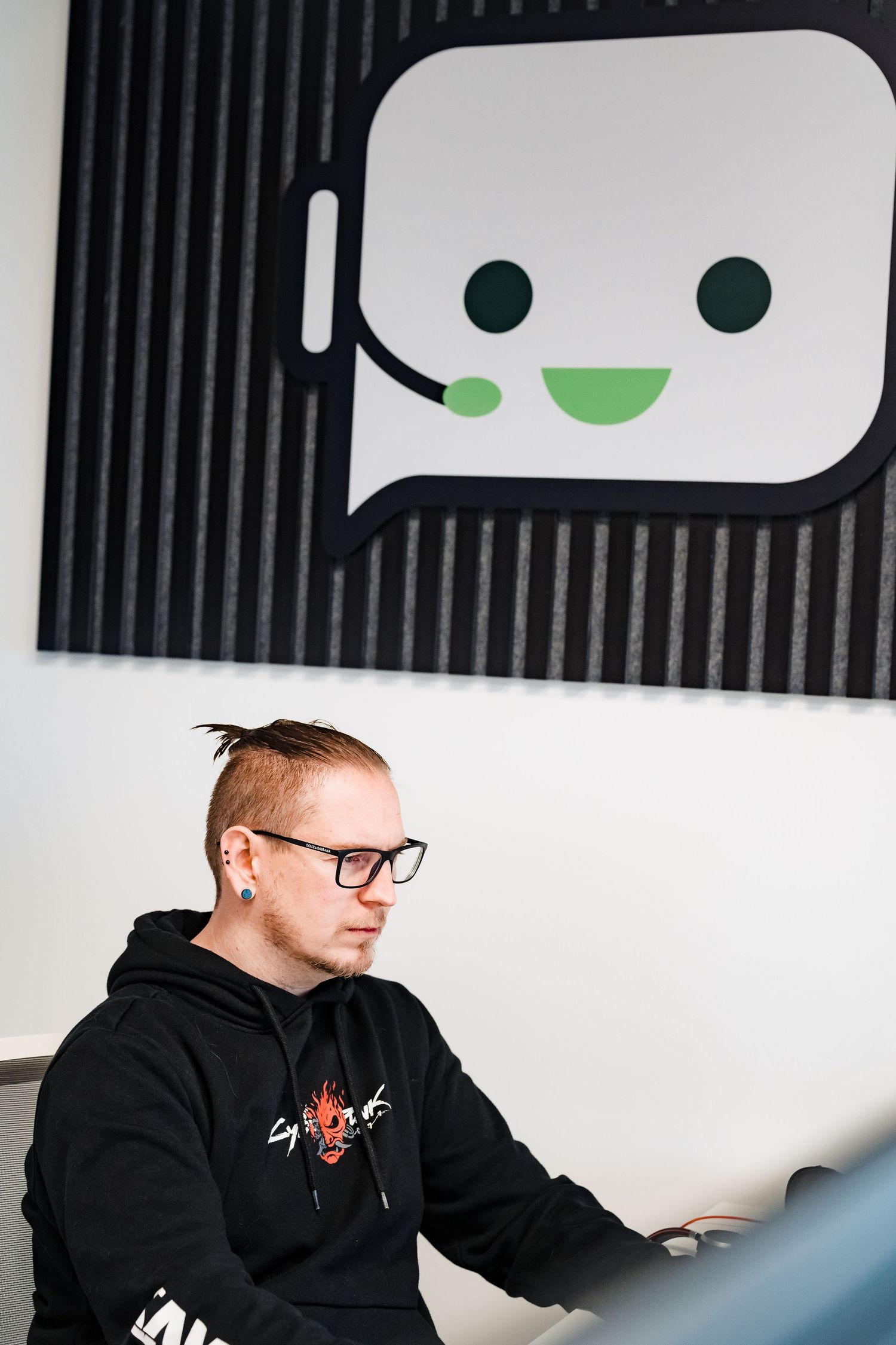 Man with glasses and a black hoodie works at a computer in front of a smiling headset chat bubble logo.
