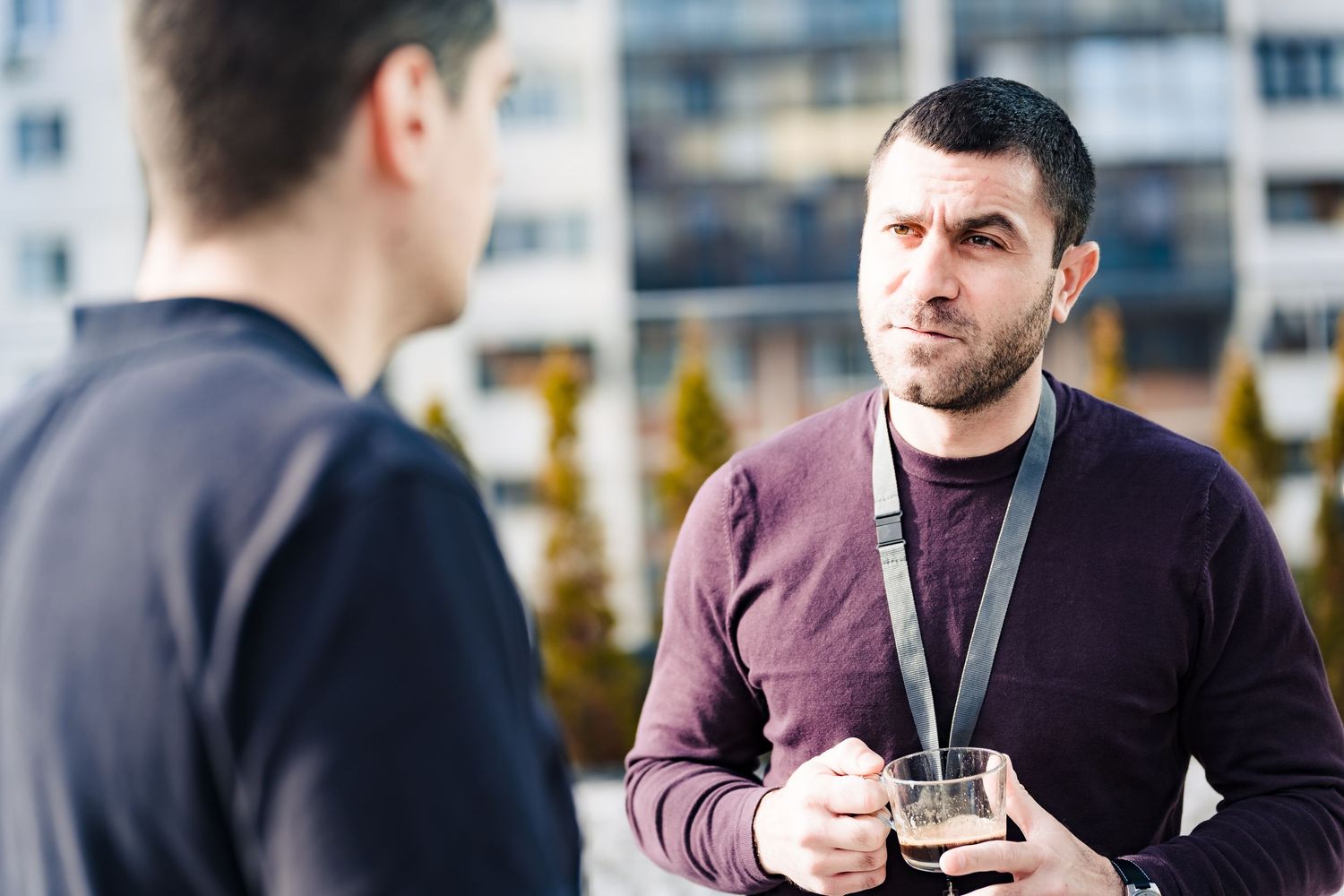 Bearded man with a lanyard holds a glass of coffee while talking with a colleague on an outdoor terrace.