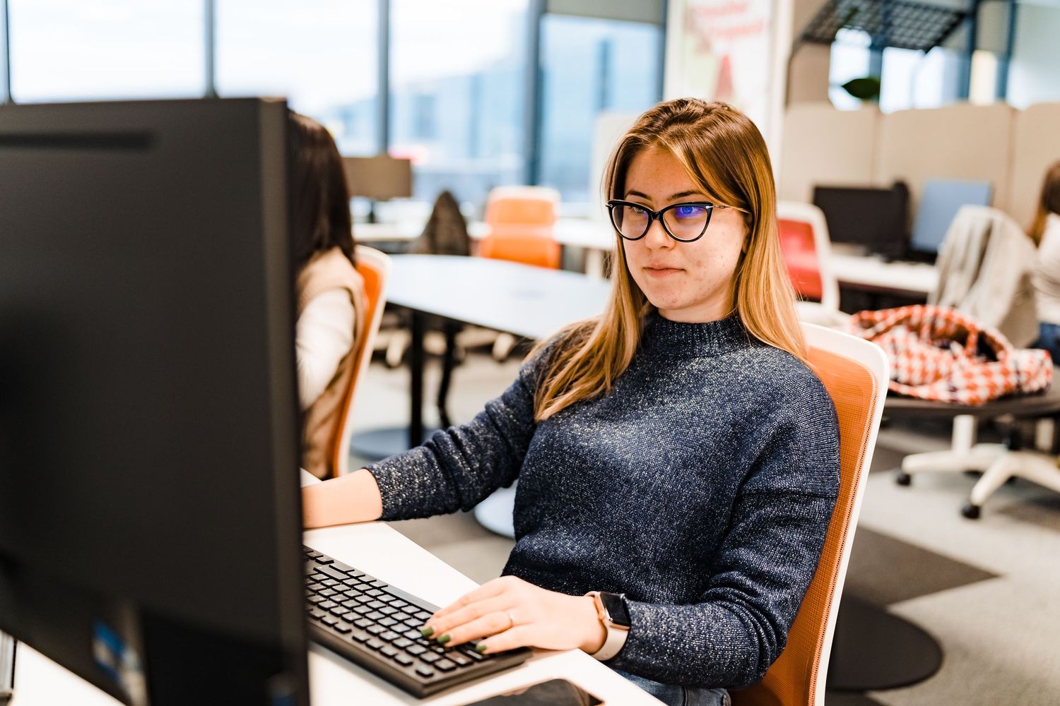 Woman with glasses and long blonde hair works at a desktop computer in a bright open-plan office with orange chairs.