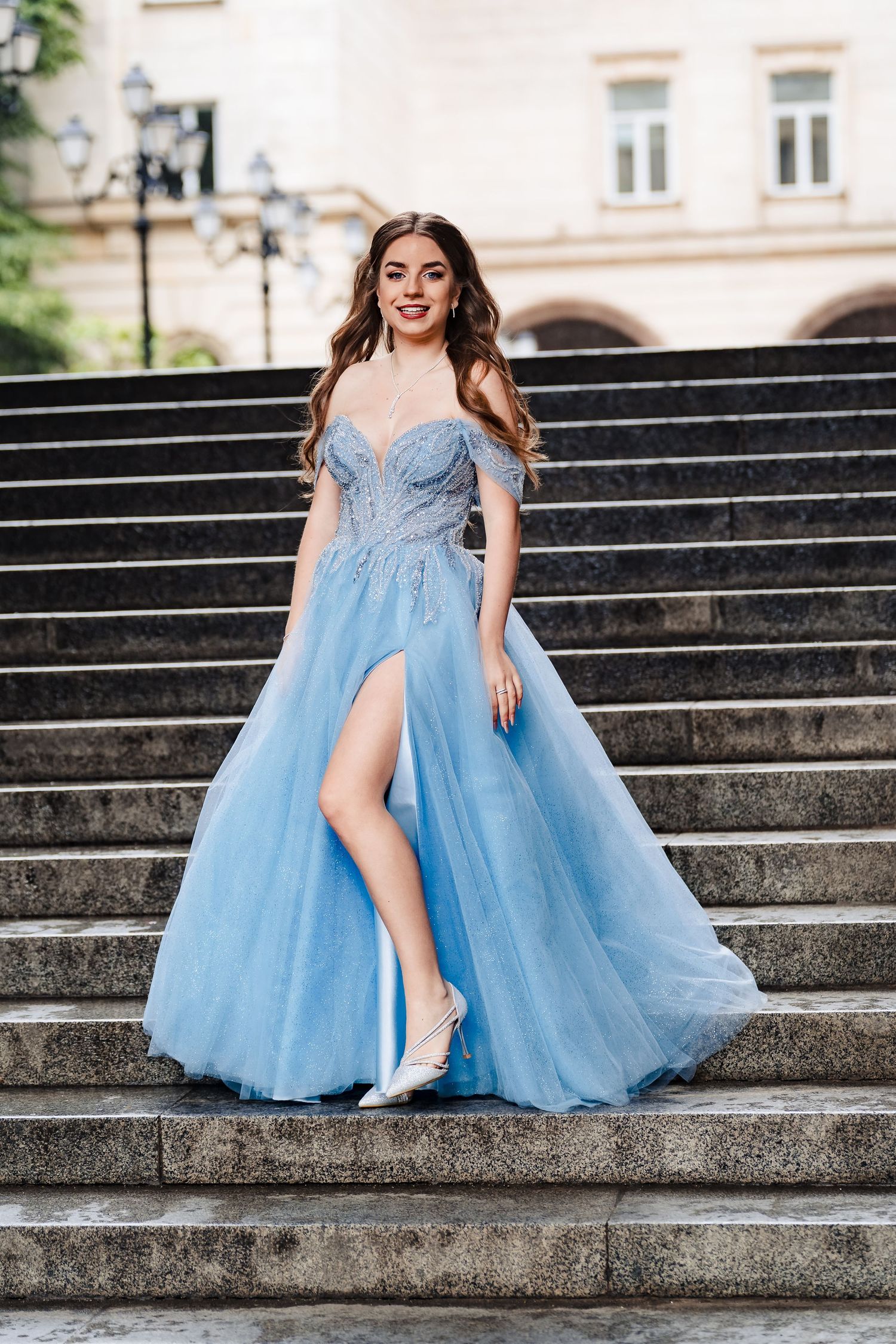 Young woman in a flowing light blue tulle gown with a high leg slit poses on stone steps in front of a historic building.