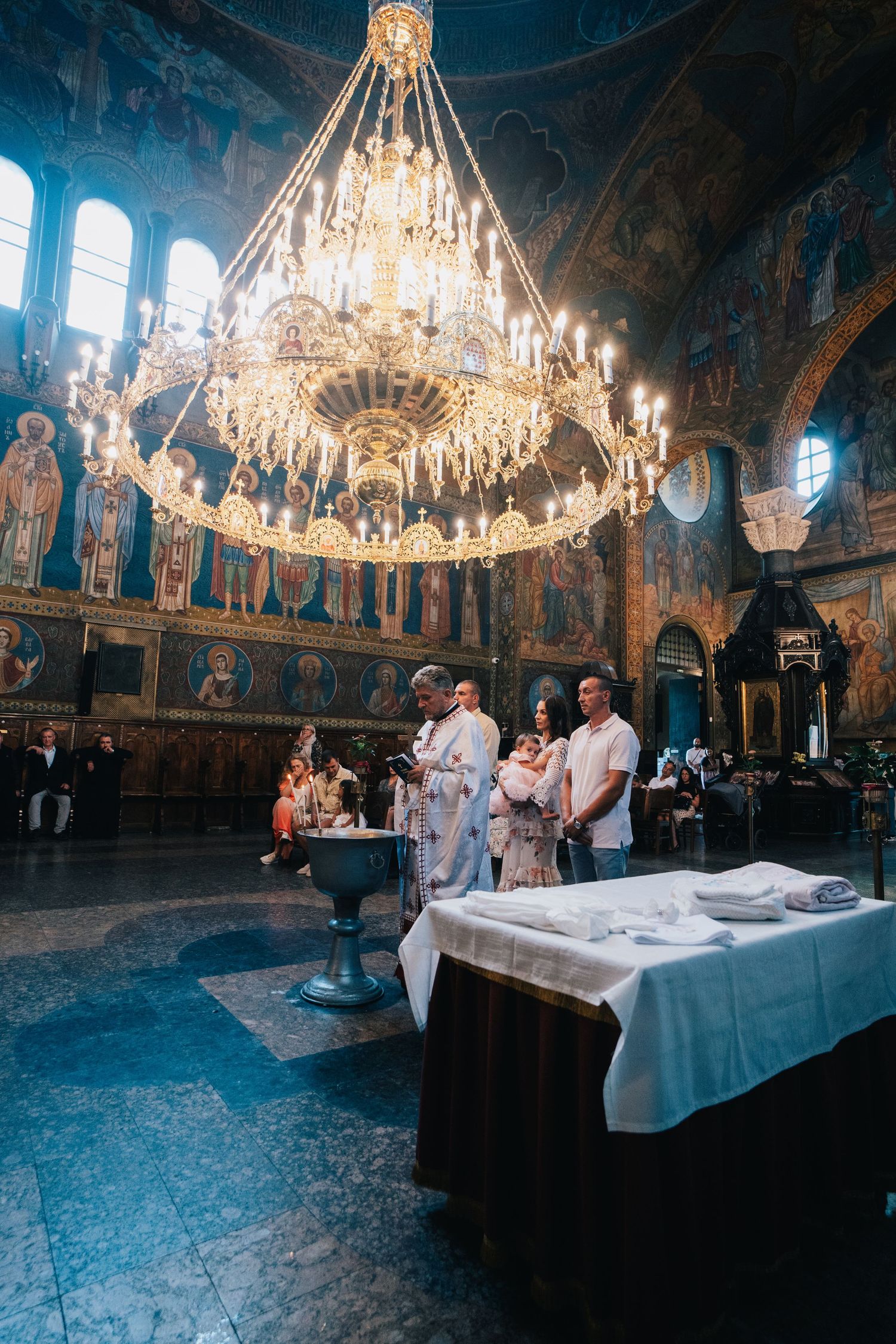 A priest leads an Orthodox baptism ceremony beneath a large ornate chandelier in a church with richly painted icon-covered walls.