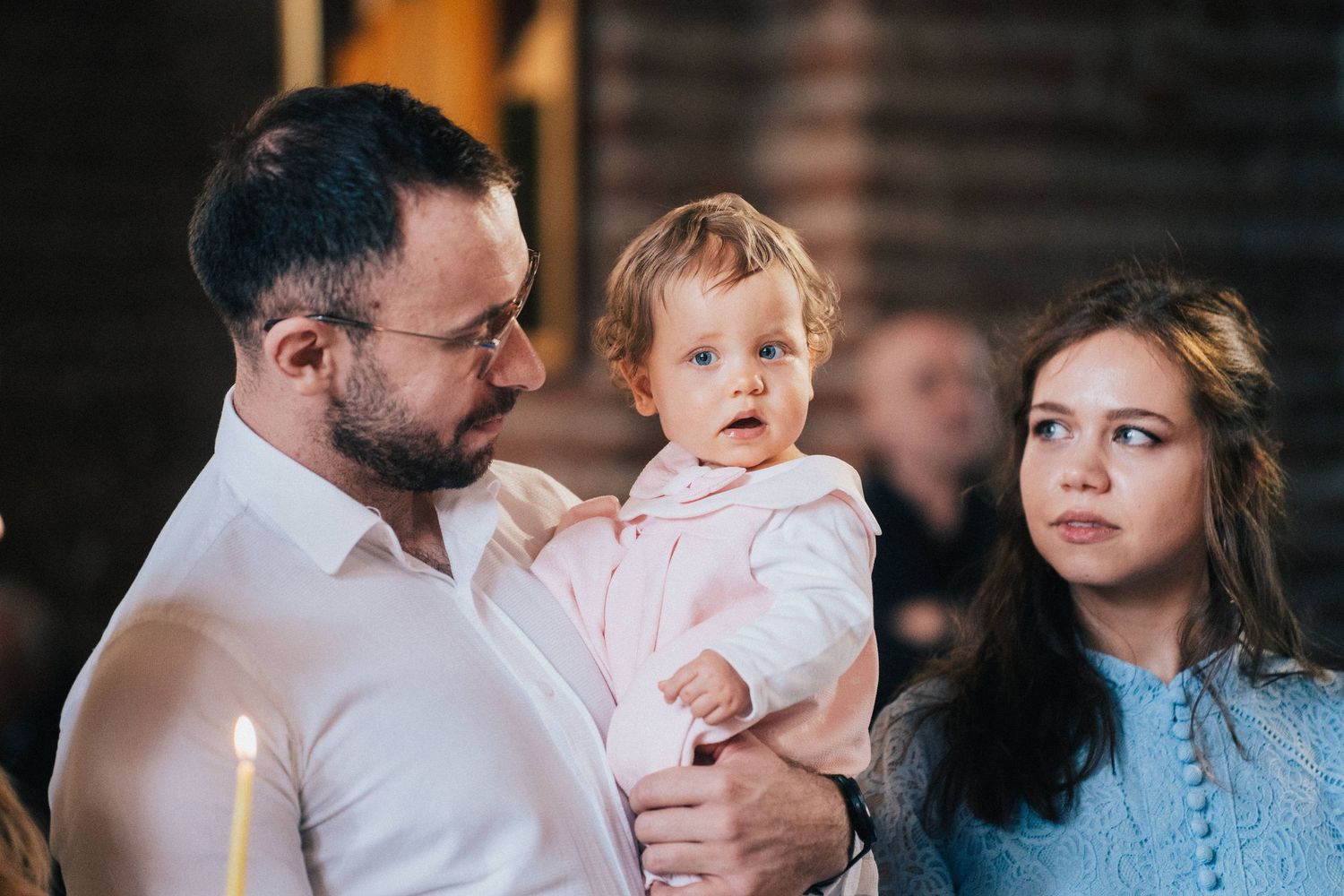A father in glasses holds his toddler daughter in pink while the mother stands beside them during a candlelit church baptism ceremony.