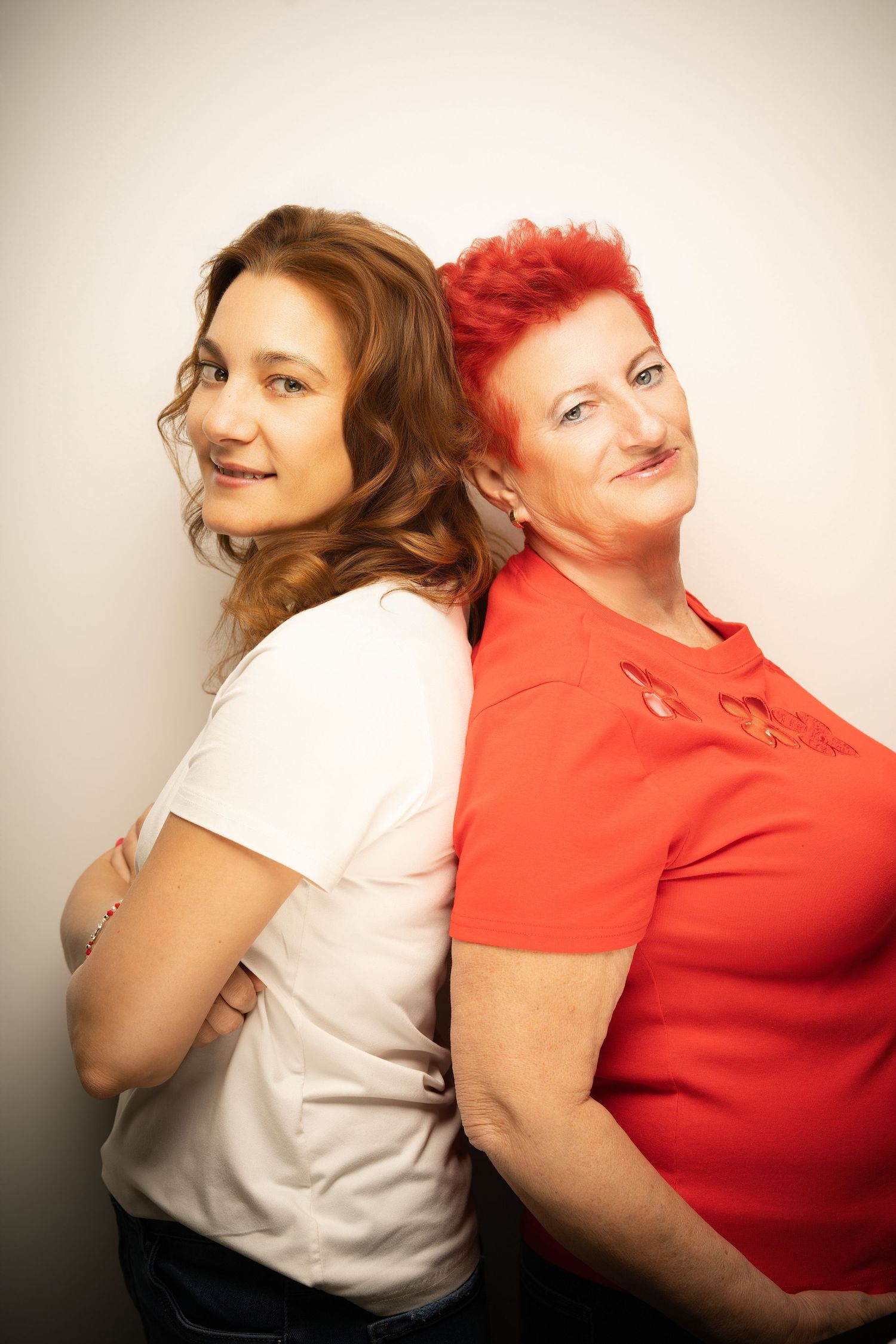 A young woman in white and a red-haired woman in a red top stand back to back smiling in a bright studio portrait.