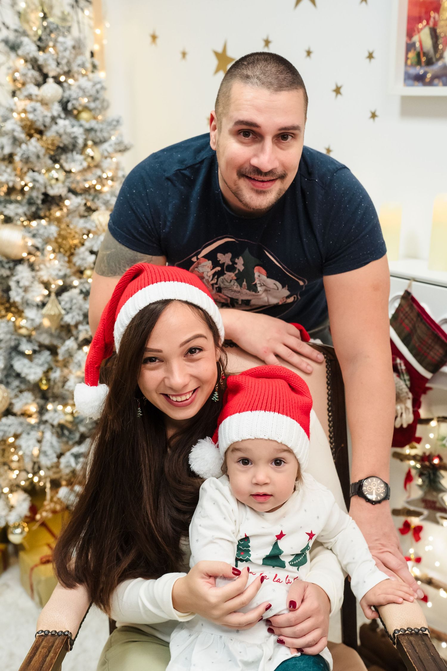 Smiling couple and baby in Santa hats pose together in front of a sparkling white Christmas tree and holiday decor.