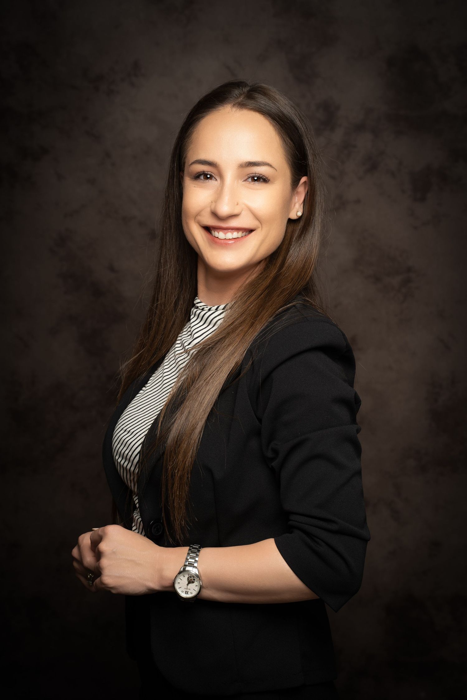 A smiling woman with long brown hair wears a black blazer over a striped top in a professional studio portrait.