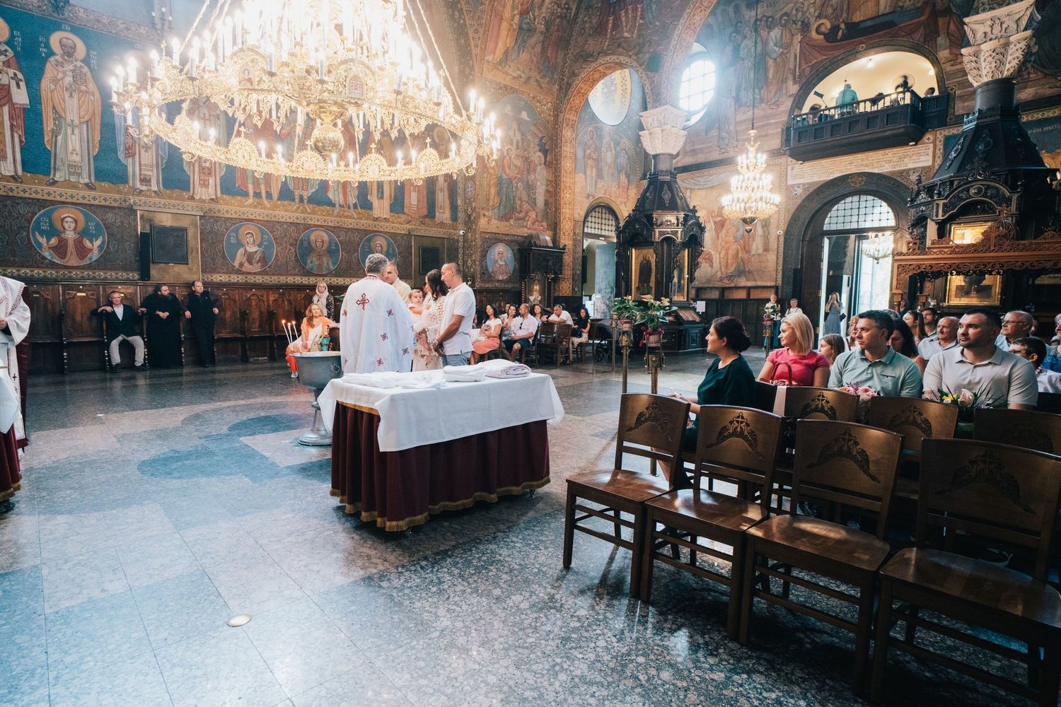 Interior of an ornate Orthodox church with a grand chandelier as a priest performs a baptism ceremony and guests sit on wooden chairs.