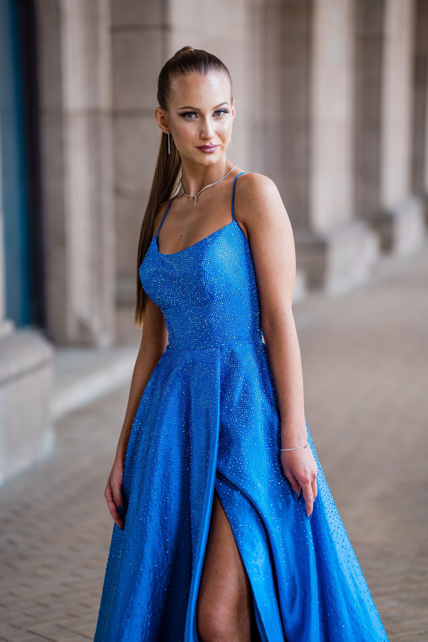 A woman with a sleek high ponytail poses in a sparkling blue gown with a high slit beside stone columns on a paved walkway.