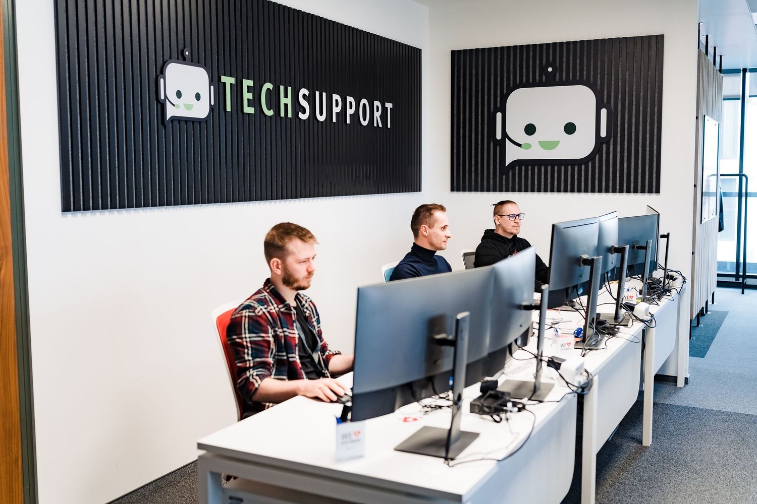 Three tech support employees work at computer desks beneath a Tech Support sign with a friendly chatbot icon.