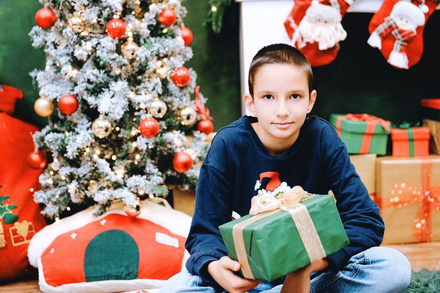 Smiling boy in a navy sweater holds a green wrapped Christmas gift beside a decorated tree, with presents and Santa stockings nearby.