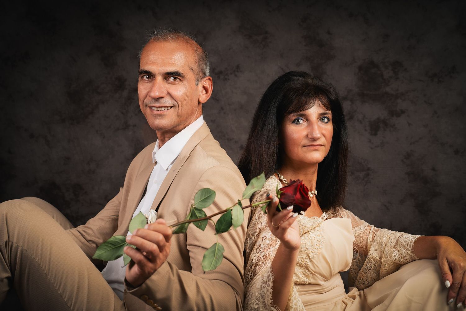 A smiling man in a beige suit and a woman in a cream lace dress sit back to back in a studio portrait, holding a red rose.