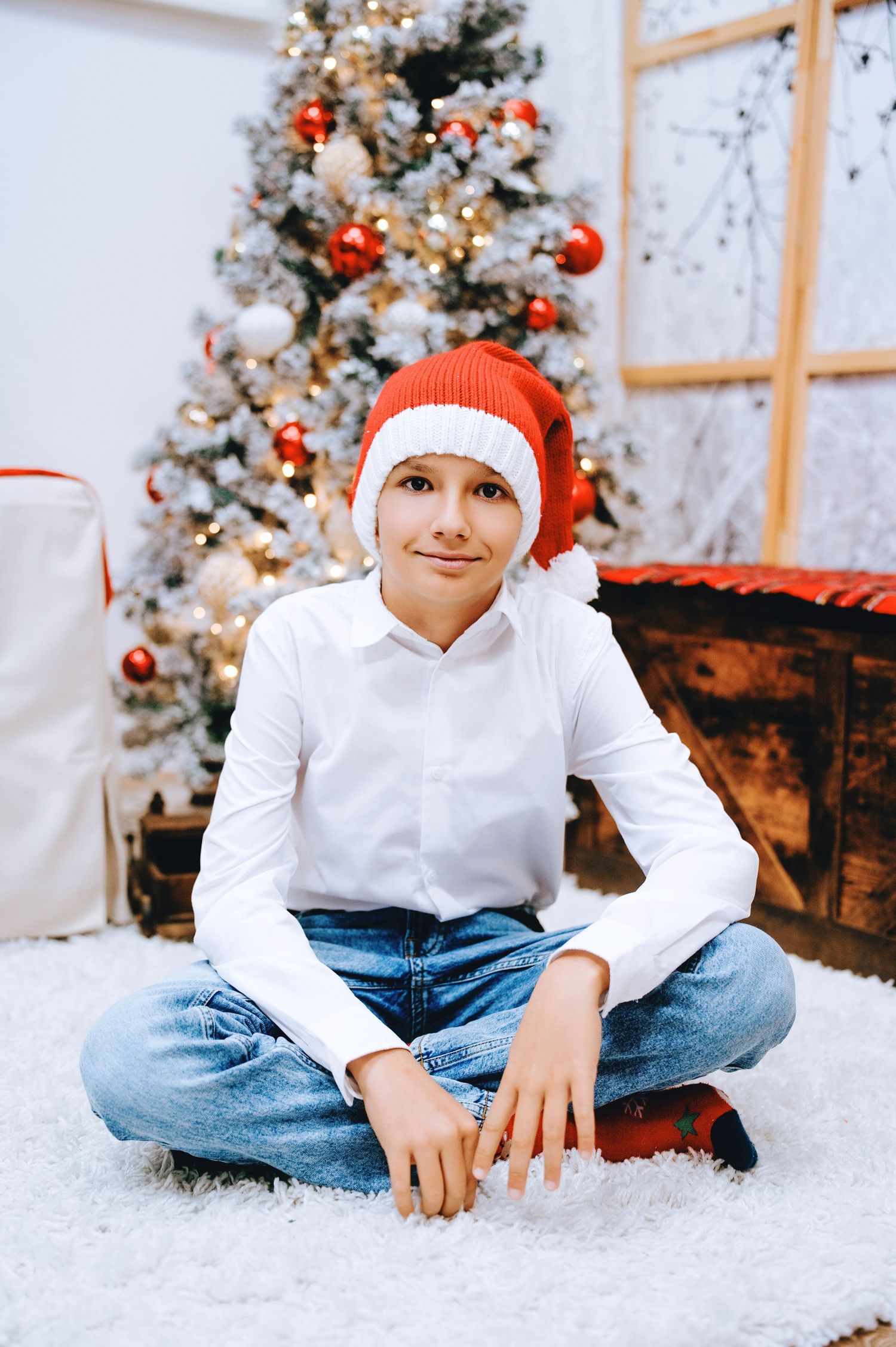 Smiling boy in a red Santa hat and white shirt sits cross-legged on a fluffy white rug beside a decorated Christmas tree.