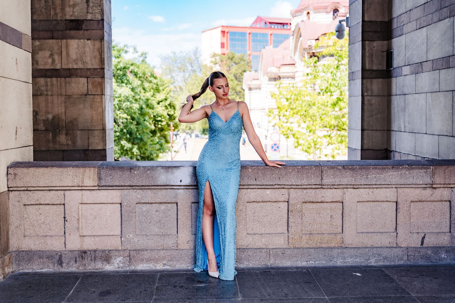 Young woman in a long blue sequin gown with a high slit poses in a stone archway overlooking a sunny city.