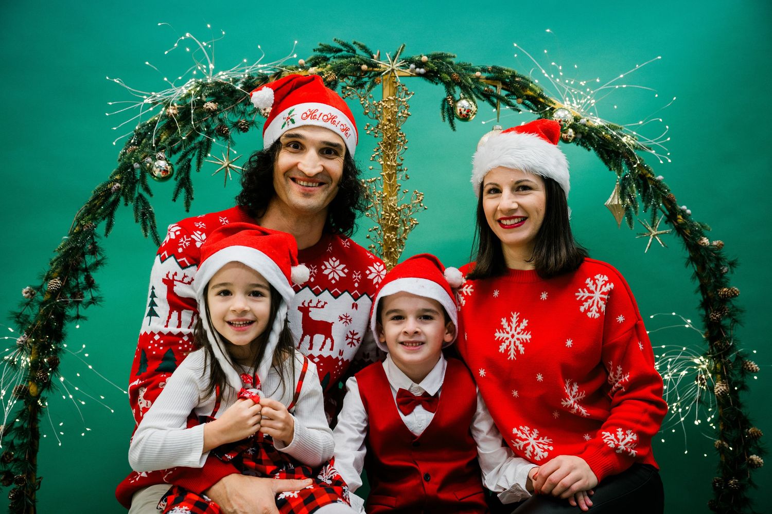 Family of four in Santa hats and festive sweaters pose with sparklers under a greenery arch against a teal backdrop.