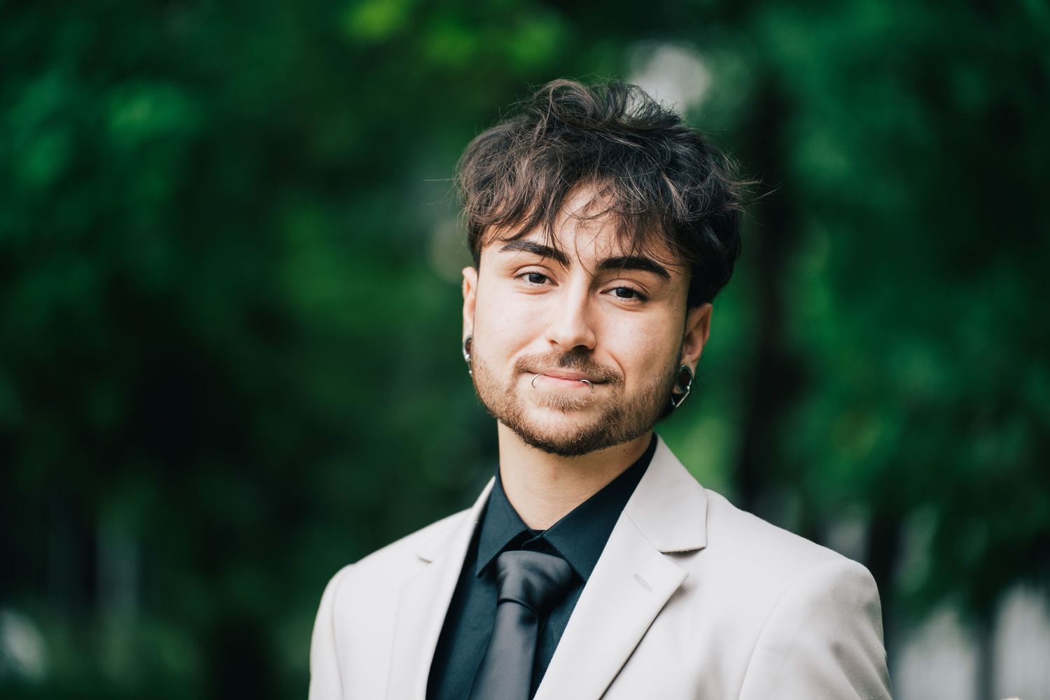 A young man with dark tousled hair and light stubble smiles in a beige blazer and dark tie, standing in front of green trees.