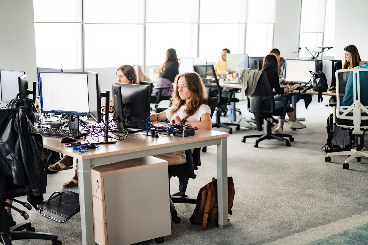 A woman types at a desk in a spacious open-plan office with rows of computer monitors and colleagues working in the background.