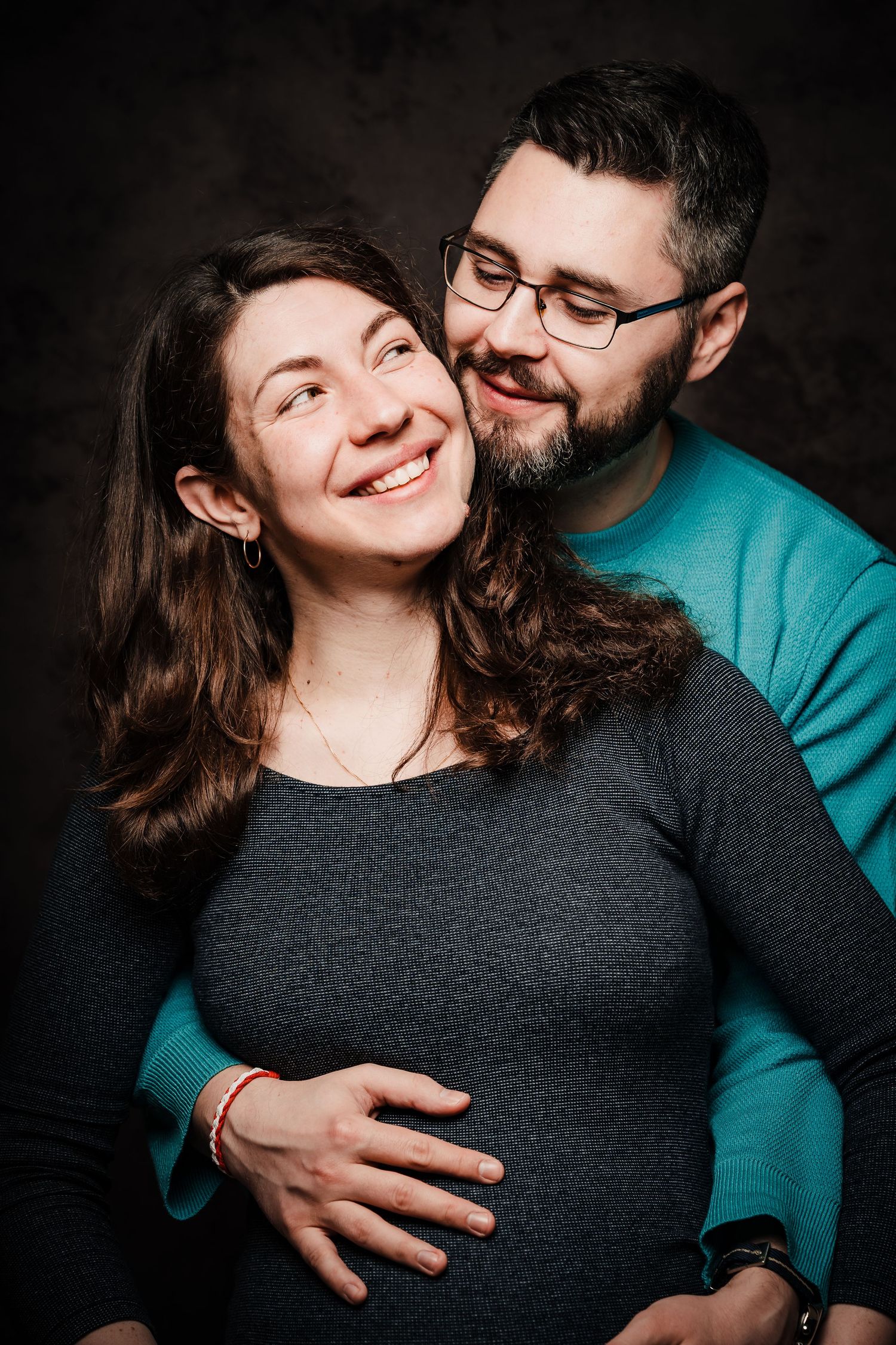 Smiling pregnant woman in a gray top embraced from behind by a bearded man in glasses wearing teal, in a studio maternity portrait.