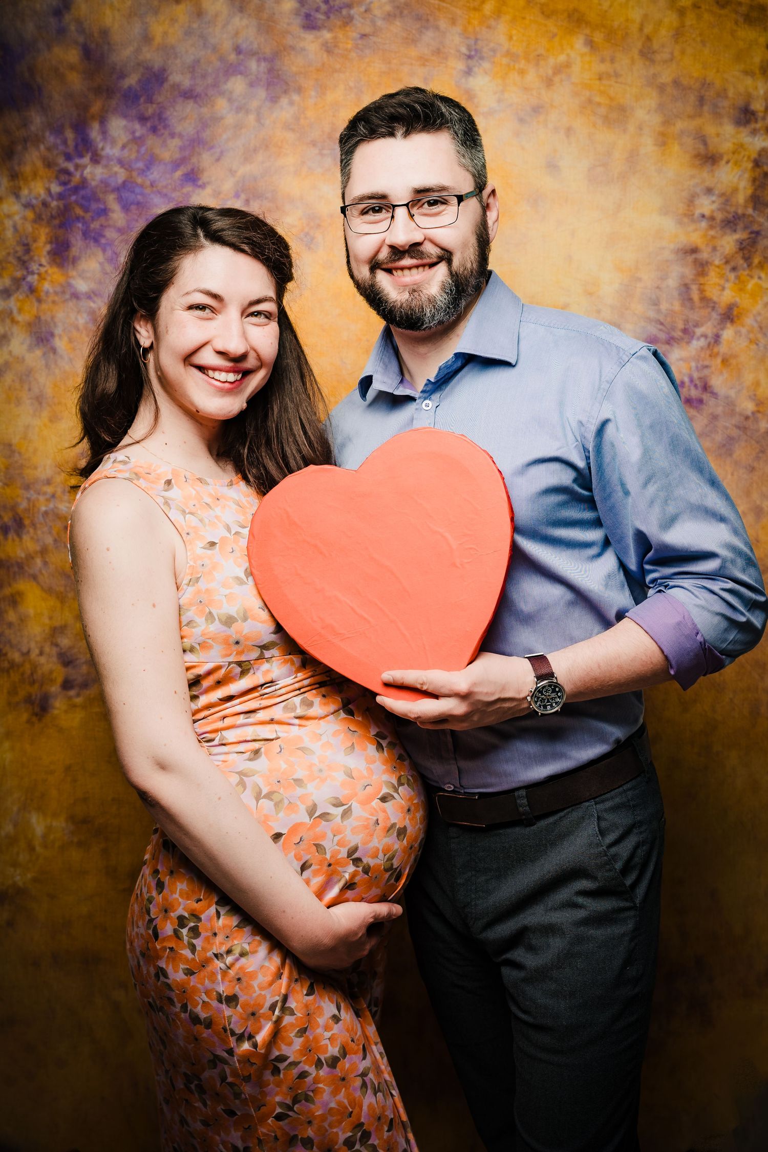 Smiling bearded man in glasses holds a red heart beside his pregnant partner in a floral dress in a maternity photo shoot.
