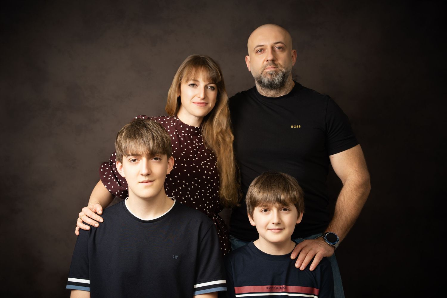 A family of four poses in a studio portrait, with parents standing behind two boys in dark t-shirts on a brown backdrop.