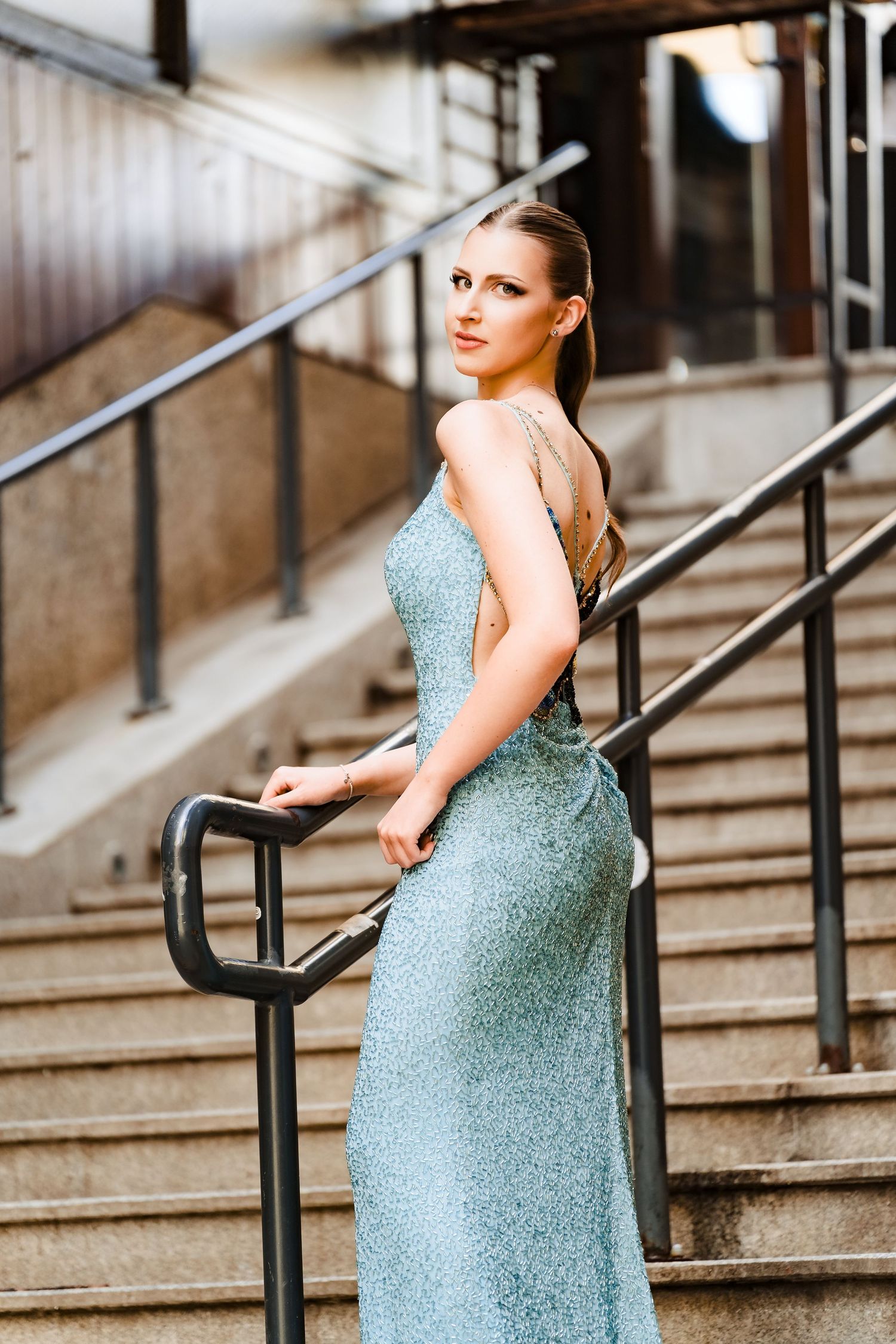 Woman in a sparkling light blue evening gown standing on outdoor stone steps with a dark metal handrail.