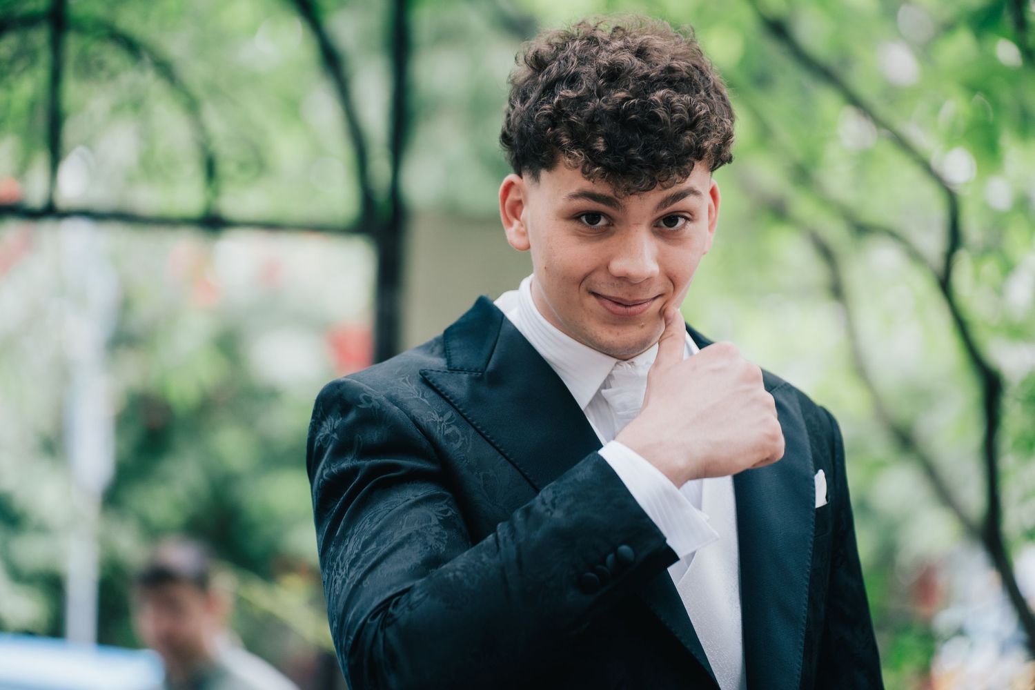 Young man with curly hair in a black formal jacket and white shirt smiles and gives a thumbs-up among green foliage.