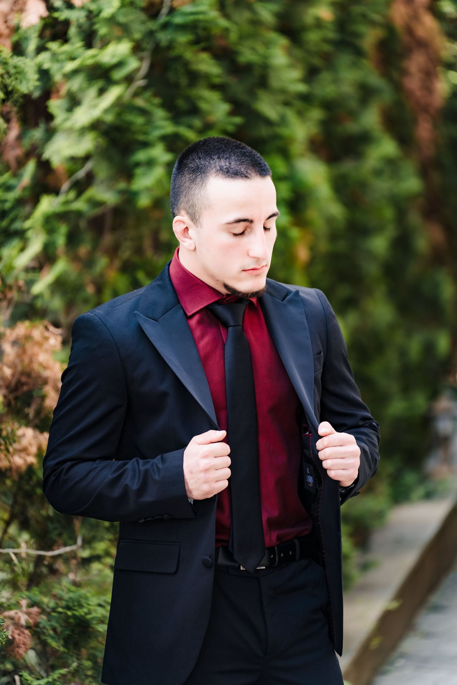 Young man in a black blazer, burgundy shirt and black tie adjusts his lapels in front of green hedges.