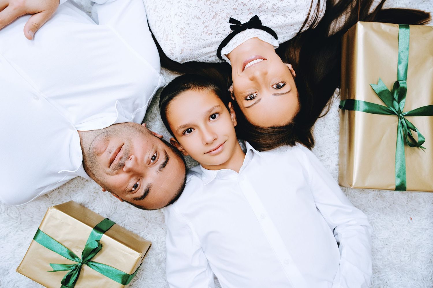 Man and two girls in white outfits lie head to head on a white fluffy rug beside gift boxes wrapped in gold paper with green ribbons.