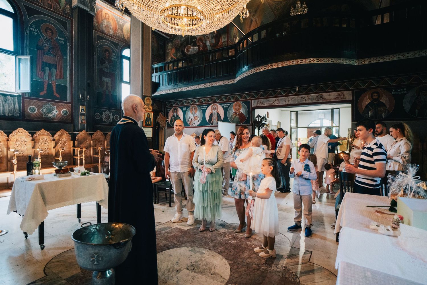 Families and children gather around a priest in a bright, ornately decorated Orthodox church before a baptism ceremony begins.