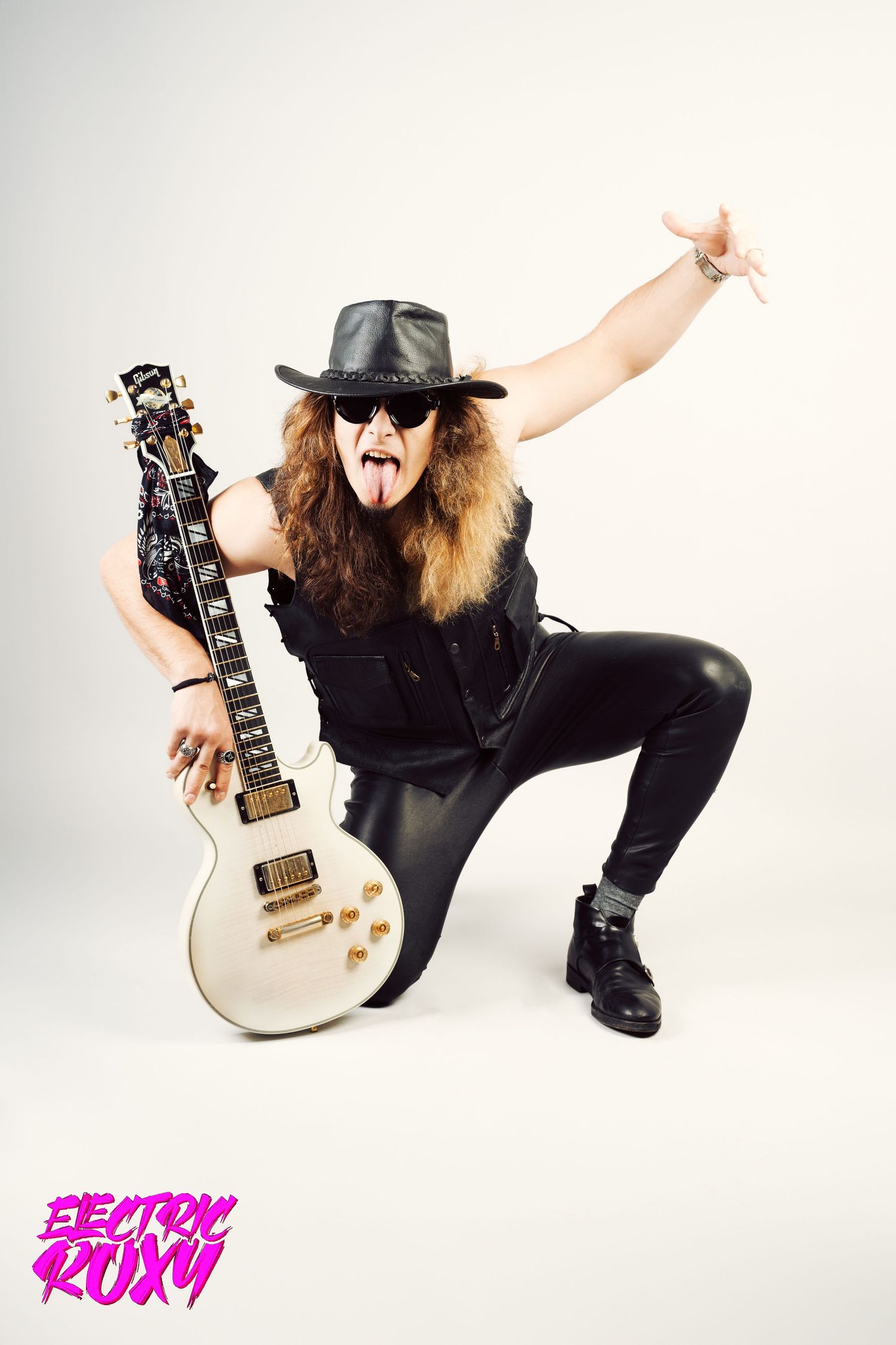 Rocker in black hat, sunglasses and leather outfit kneels with a white electric guitar, tongue out and flashing a rock hand sign.
