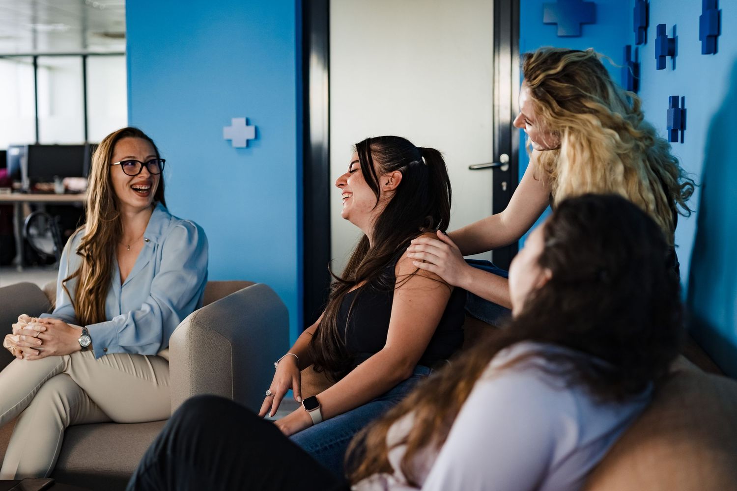 Three smiling women talk casually on lounge sofas in a blue office space with cross-shaped decorations on the wall.