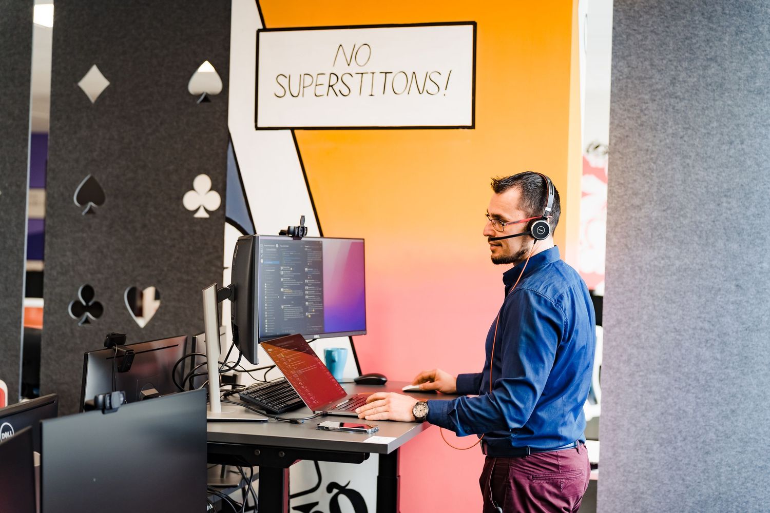 Man wearing a headset works at a standing desk with monitors beside a colorful wall sign reading No Superstitions.