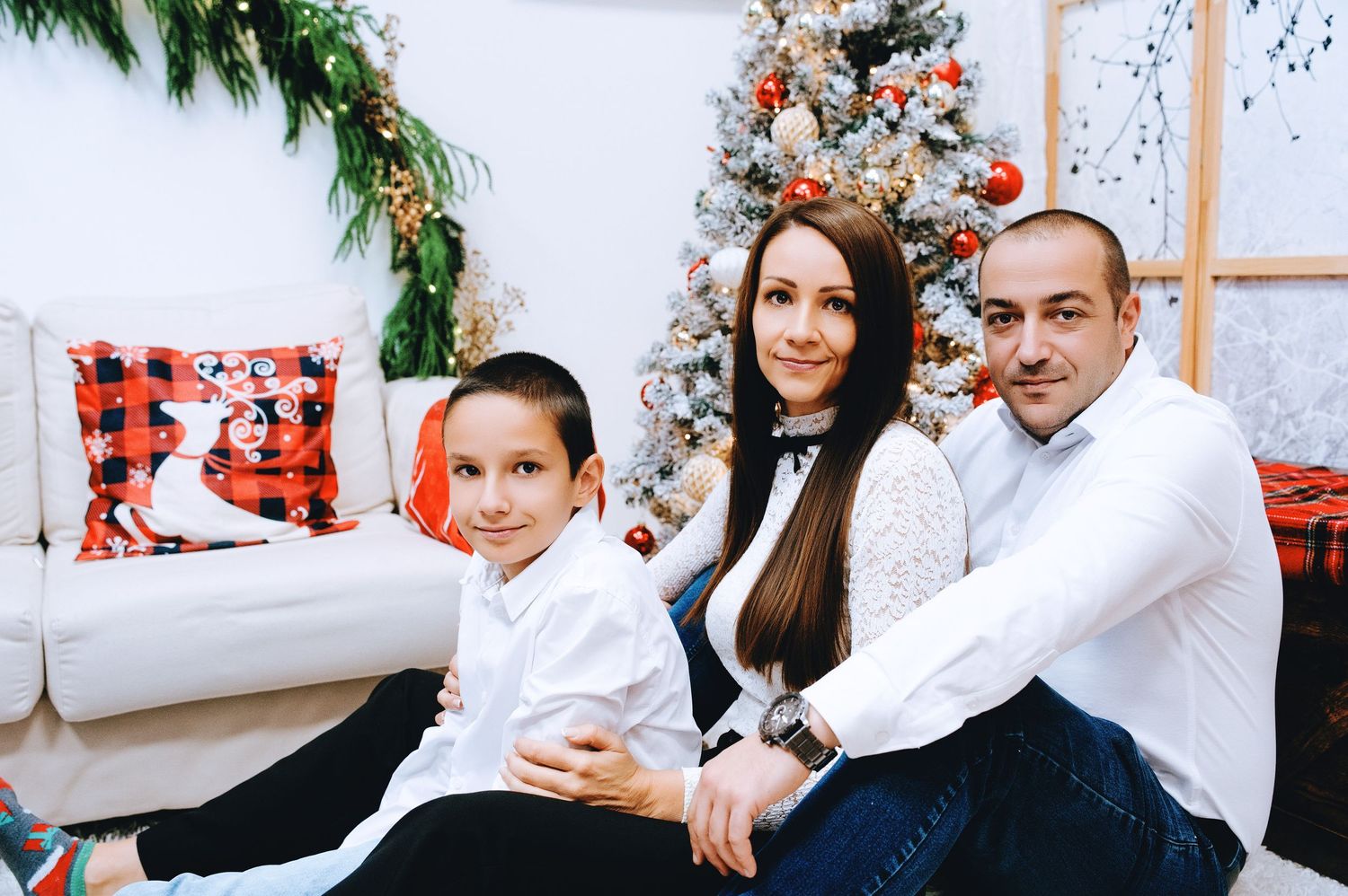 Smiling family of three in white shirts poses on a couch beside a decorated Christmas tree with red ornaments and festive plaid pillows.