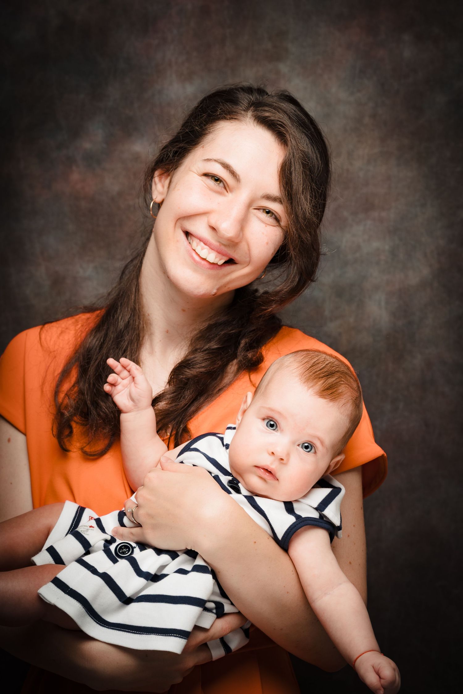 A smiling mother in an orange dress cradles her baby in a striped outfit against a textured brown studio background.