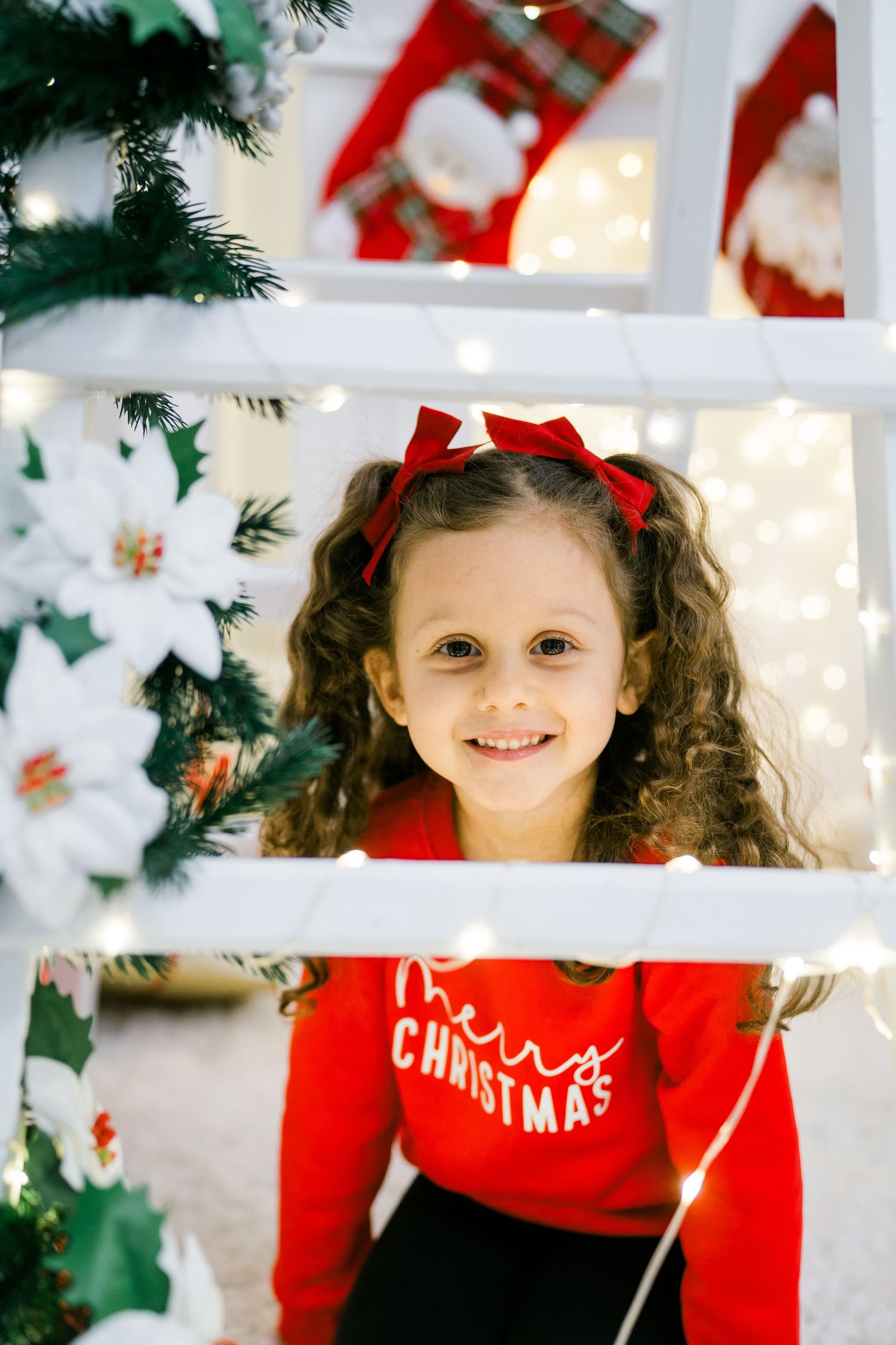 Smiling little girl with a red hair bow wearing a Merry Christmas sweatshirt beside a white ladder and festive fairy lights.