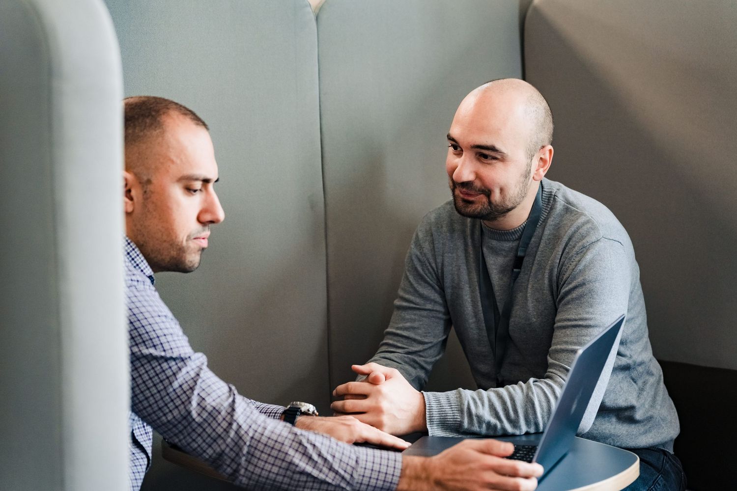 Two men sit in a quiet office nook having a focused conversation over an open laptop.