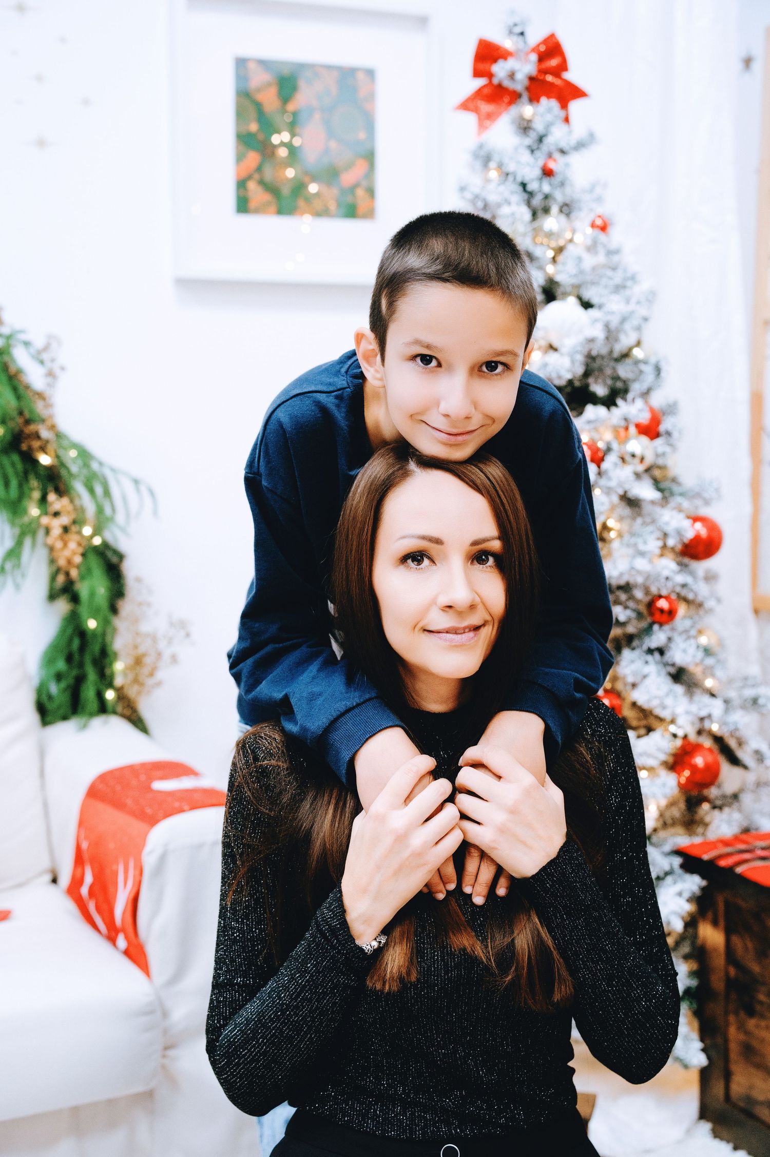 Smiling boy in a navy sweater leans over a woman with long dark hair beside a Christmas tree with red ornaments and a red bow.