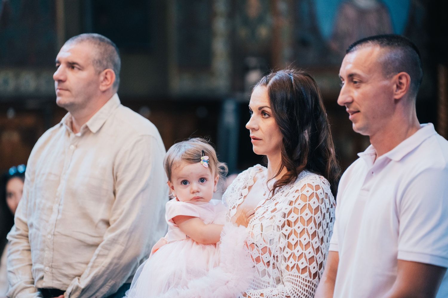 Parents and a toddler in a white dress stand together during an Orthodox baptism ceremony inside a church.