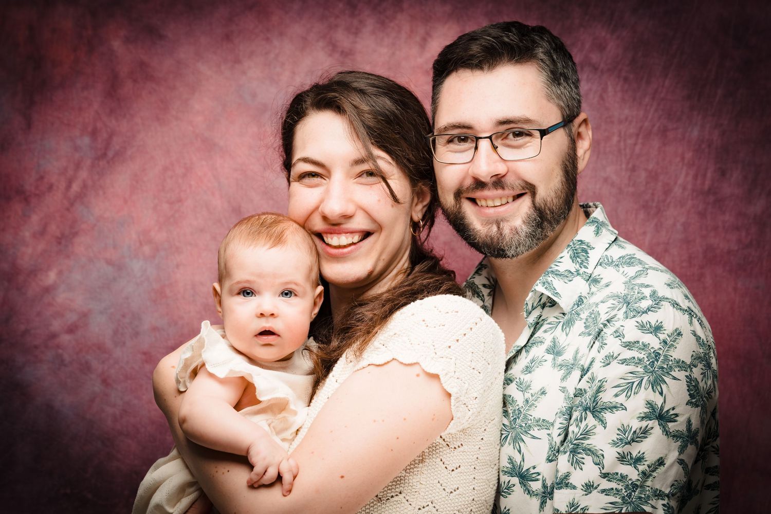Smiling woman and bearded man in glasses pose cheerfully with their baby in a warm family portrait against a mottled pink studio backdrop.