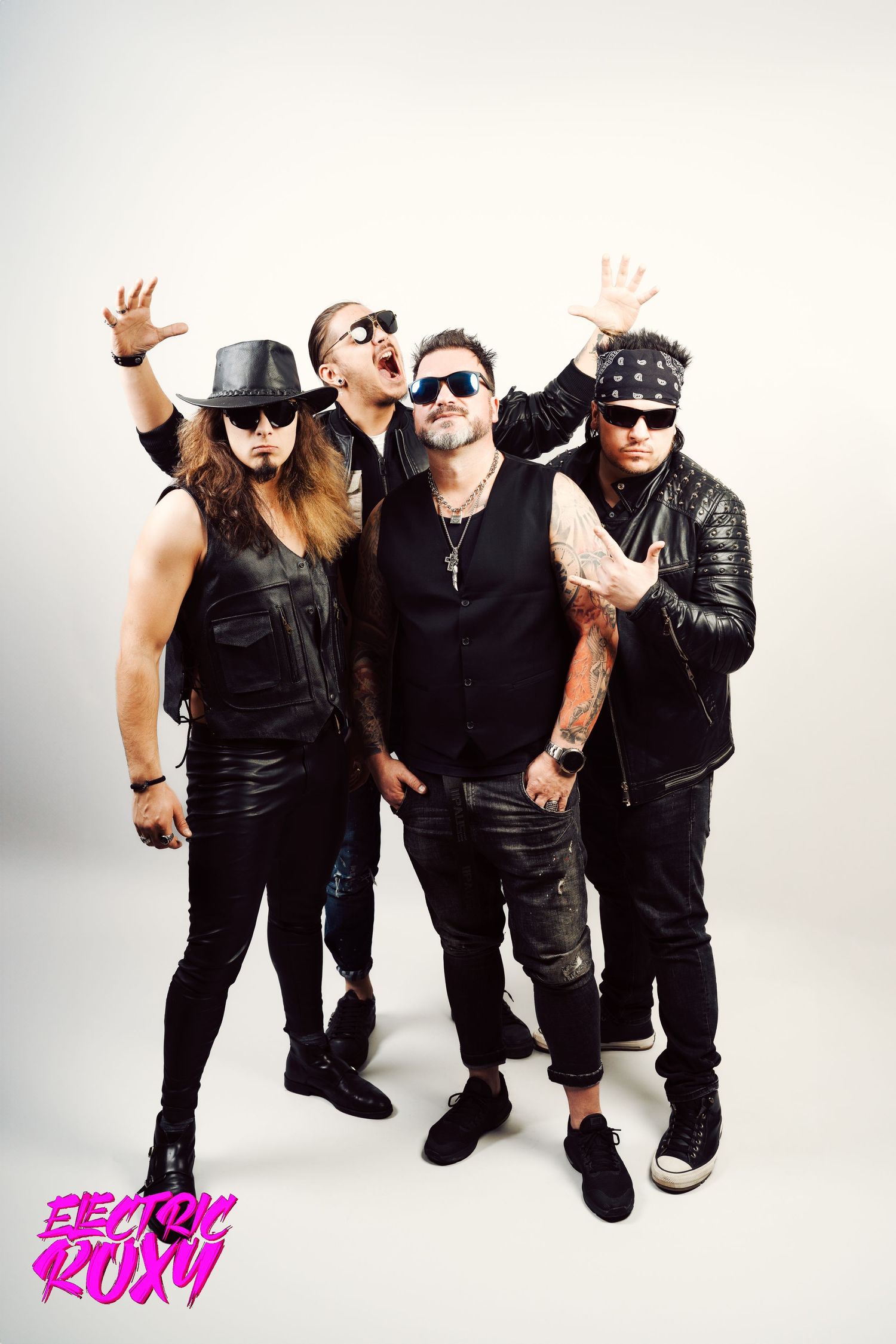 Four rock musicians in black leather, sunglasses and a hat pose with raised hands against a white backdrop with an Electric Roxy logo.