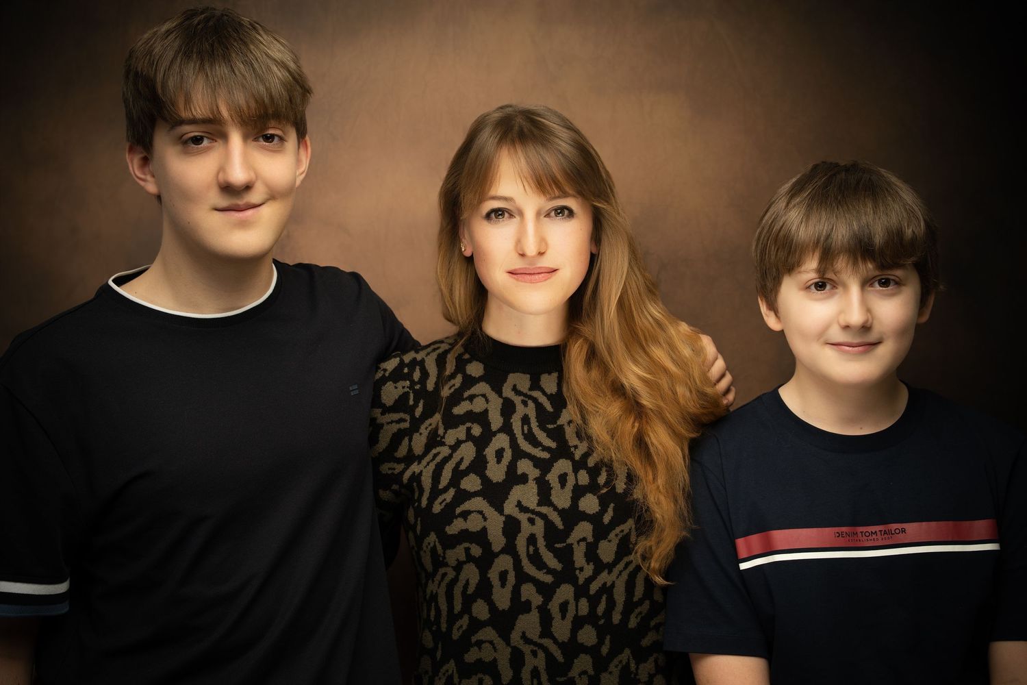 Studio family portrait of three young siblings standing close together against a warm brown backdrop, smiling in dark casual clothing.