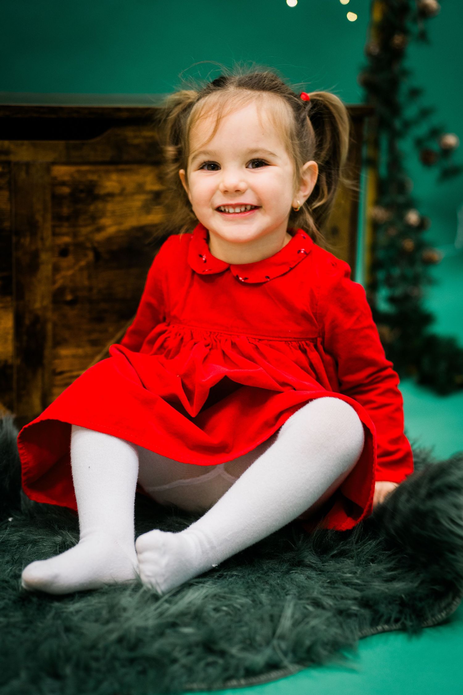 Smiling toddler girl in a red dress and white tights sits on a green fur rug beside a wooden crate and Christmas garland.