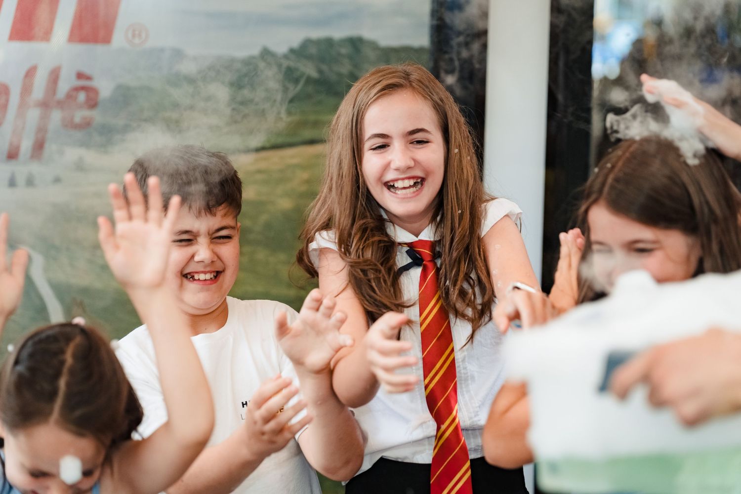 Smiling children in school uniforms laugh and wave their hands while playing together outdoors on a sunny day.