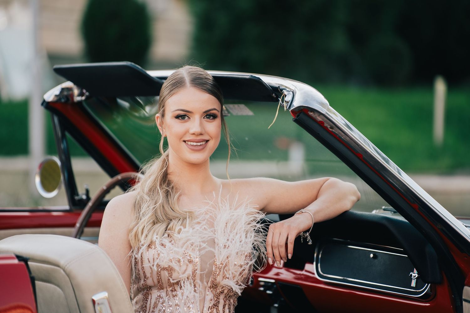 Young woman in a feathered blush sequin dress smiles while leaning on the door of a red vintage convertible.